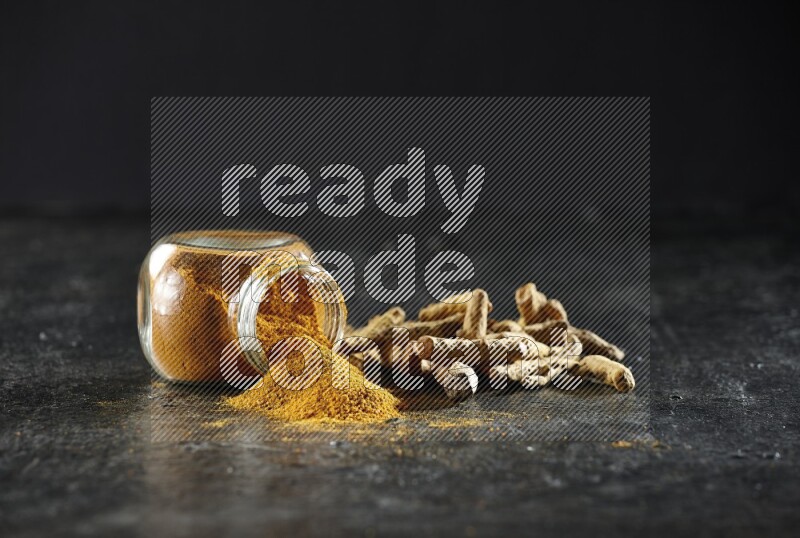 A flipped glass spice jar full of turmeric powder and powder spilled out of it with dried whole fingers on textured black flooring
