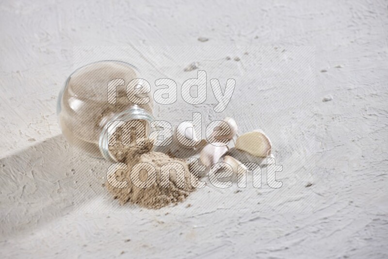 A glass spice jar full of garlic powder flipped over with the powder came out on a textured white flooring