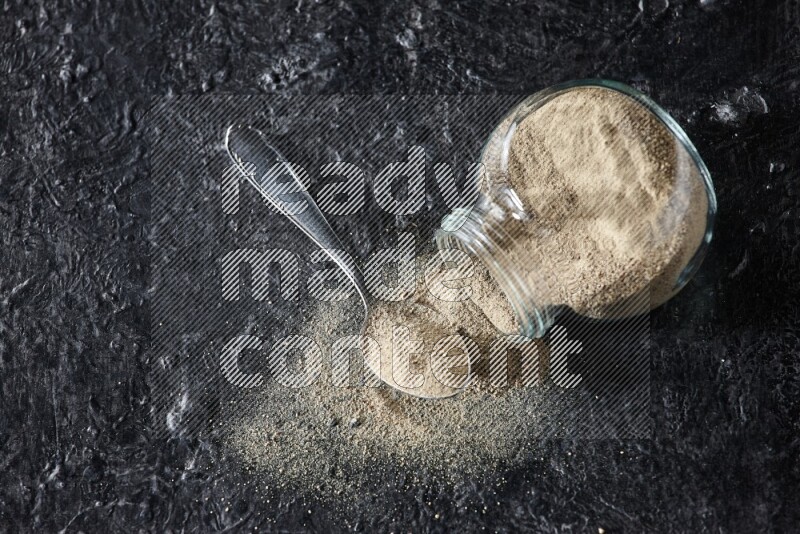 A flipped herbal glass jar and a metal spoon full of white pepper powder with spilled powder on textured black flooring