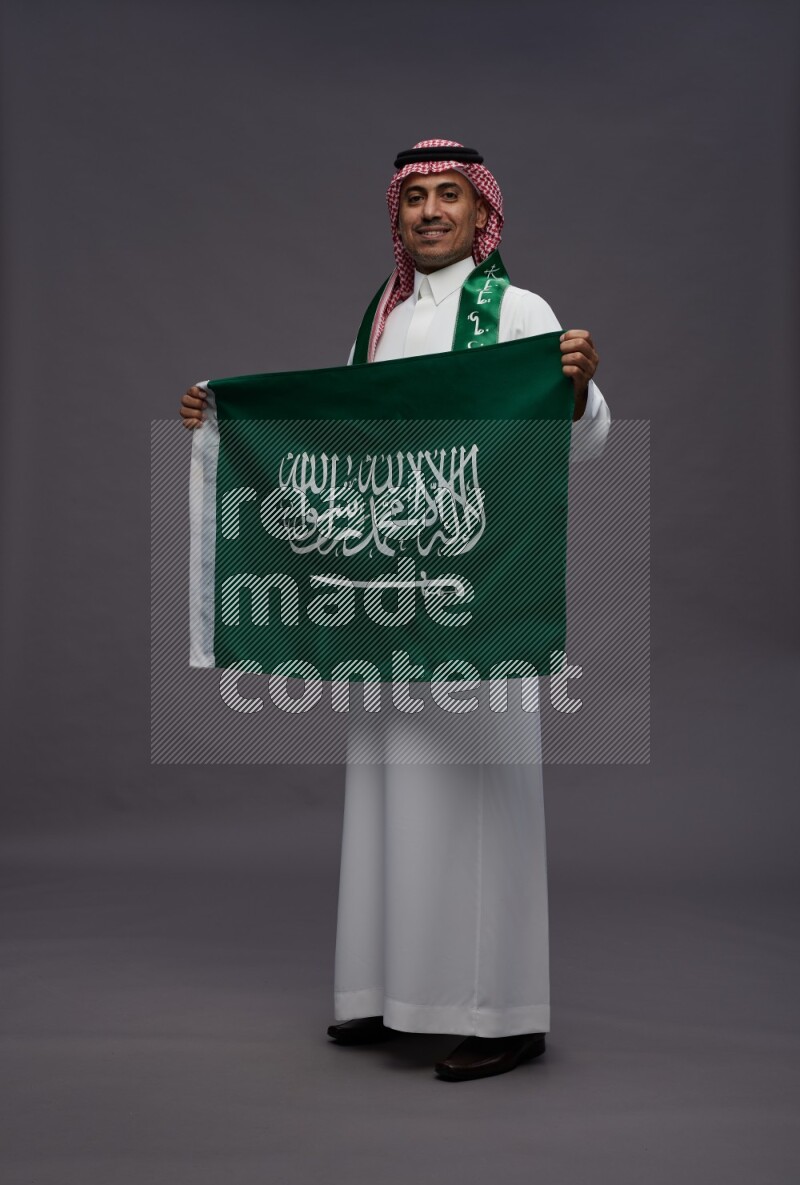 Saudi man wearing thob and shomag standing holding Saudi flag on gray background