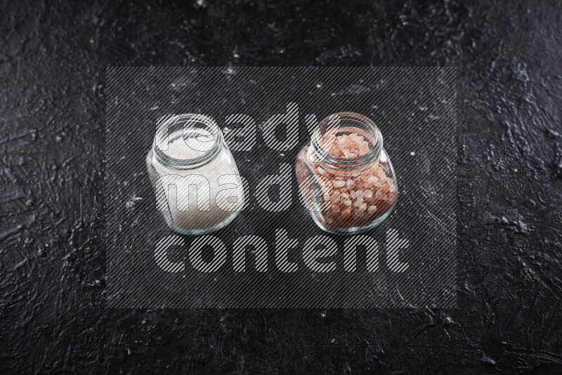 2 وعاء زجاجي احدهمها ممتلئ بملح الهيمالايا الخشن والأخر بملح البحر الخشن على خلفية سوداء