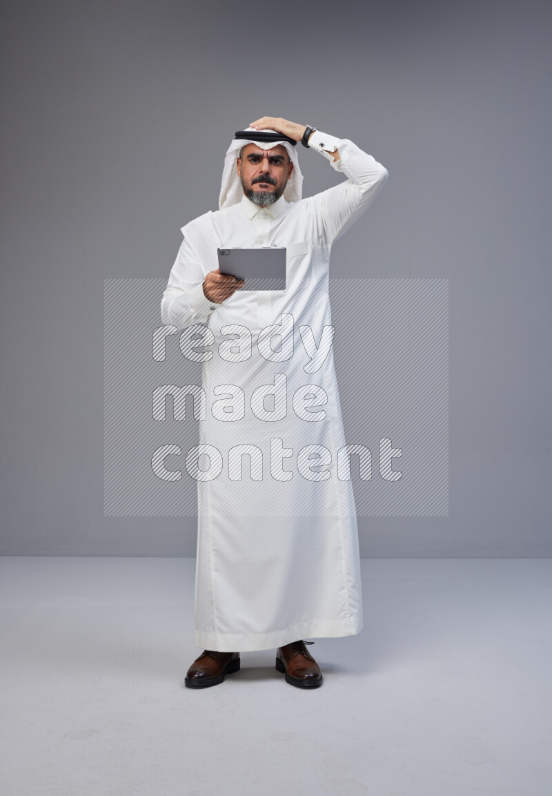 Saudi man Wearing Thob and white Shomag standing working on tablet on Gray background