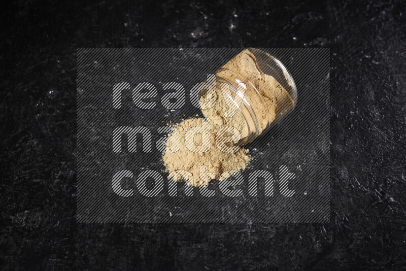 A glass jar full of ground ginger powder flipped with some spilling powder on black background