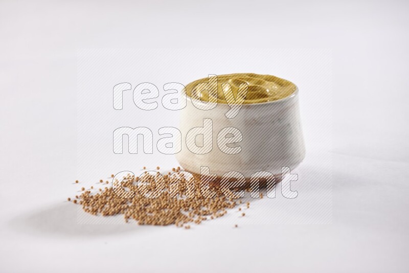 A beige pottery bowl full of mustard paste with mustard seeds underneath on white flooring