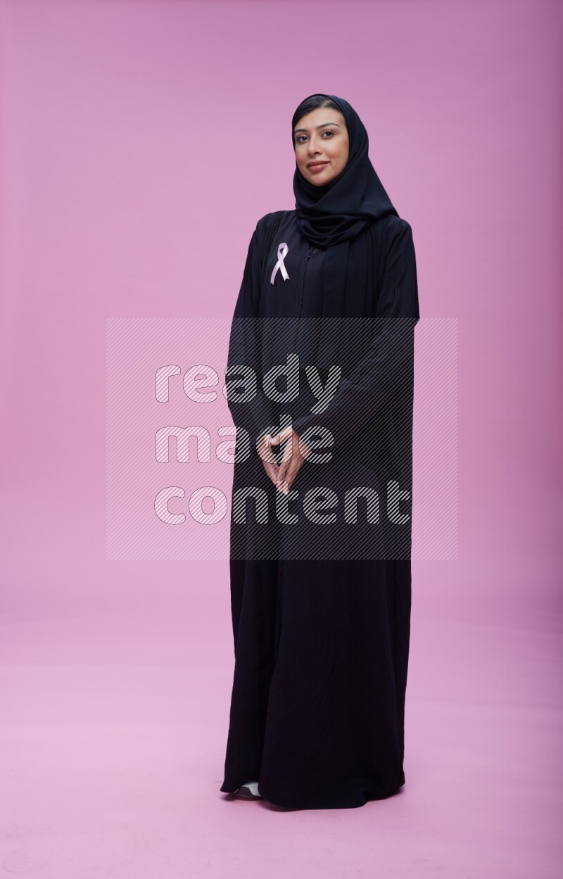 Saudi woman wearing pink ribbon on Abaya standing interacting with the camera on pink background
