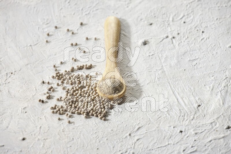 A wooden spoon full of white pepper powder and white pepper beads on textured white flooring