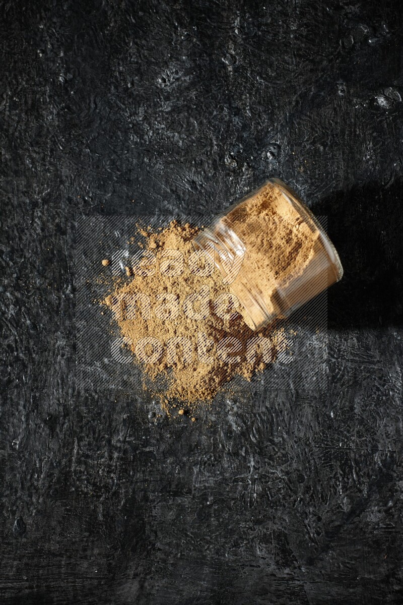 A flipped glass jar full of allspice powder and powder spilled out of it on a textured black flooring