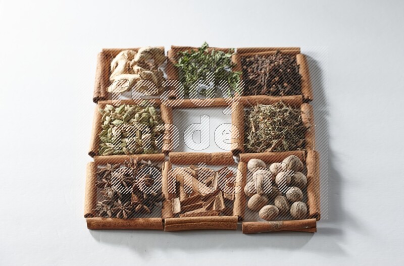 9 squares of cinnamon sticks, the middle square is empty and surrounded with dried mint, dried ginger, cardamom, star anise, cinnamon, nutmeg, dried basil and cloves on white flooring