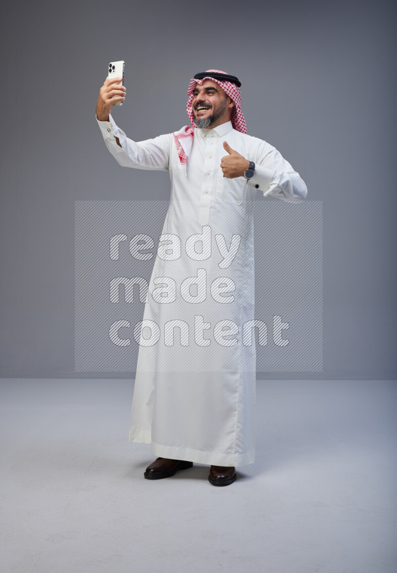Saudi man Wearing Thob and red Shomag standing taking selfie on Gray background