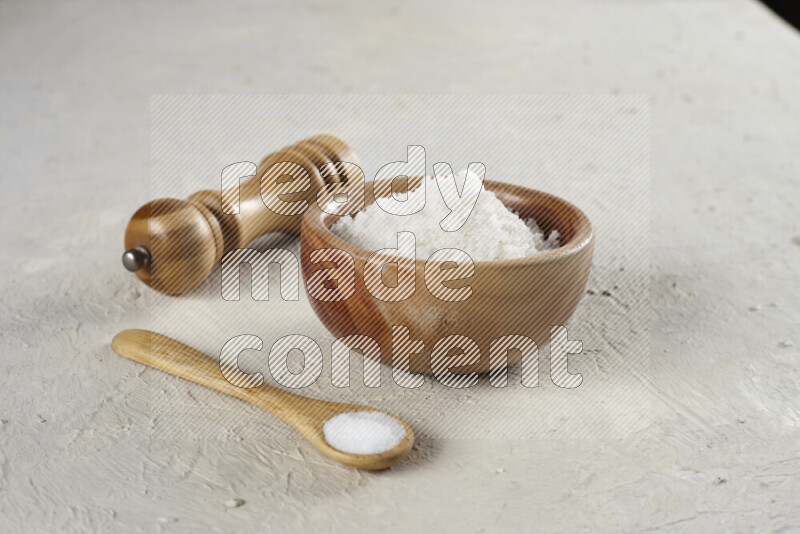 A wooden bowl and spoon filled with white sea salt and wooden grinder beside them on white background