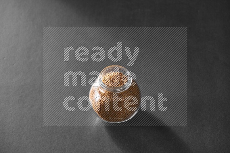 A glass spice jar full of mustard seeds on black flooring