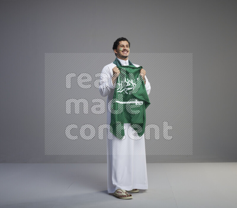 A Saudi man standing wearing thob with face painting wrapping Saudi flag scarf and holding big Saudi flag on gray background