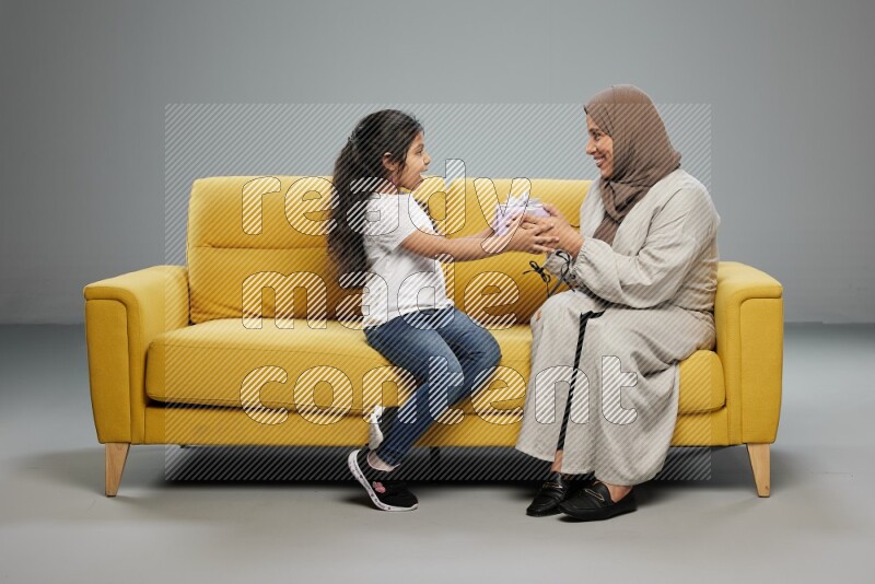 A girl sitting on a yellow sofa and giving a gift to her mother on gray background