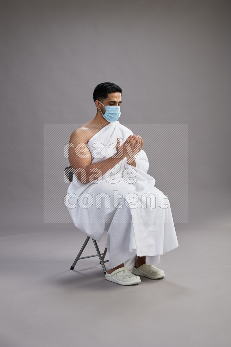 A man wearing Ehram with face mask sitting on chair dua'a on gray background