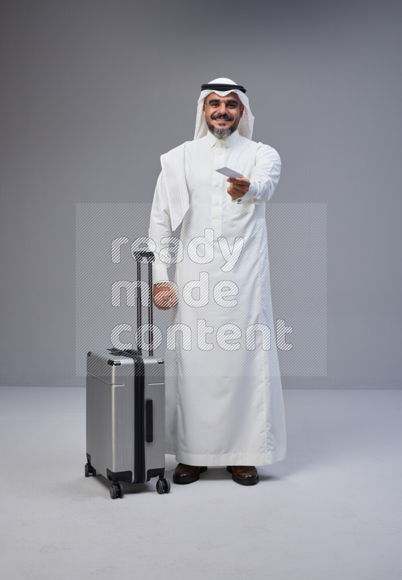 Saudi man wearing Thob and white Shomag standing holding Travel bag and ATM card on Gray background