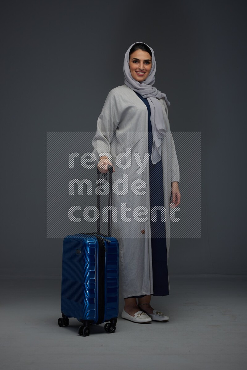 A Saudi woman wearing a light gray Abaya and head scarf standing and showing the tablet's screen eye level on a grey background