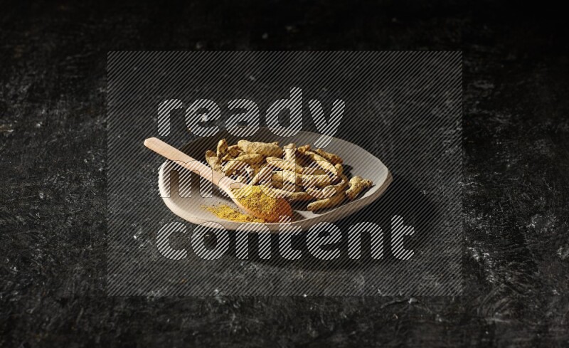 A plate filled with dried turmeric fingers and a wooden spoon full of turmeric powder on a textured black flooring