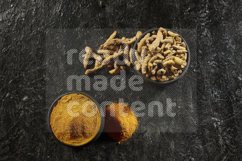 2 pottery black bowls, one full of turmeric powder and the other full of dried turmeric whole fingers on a textured black flooring
