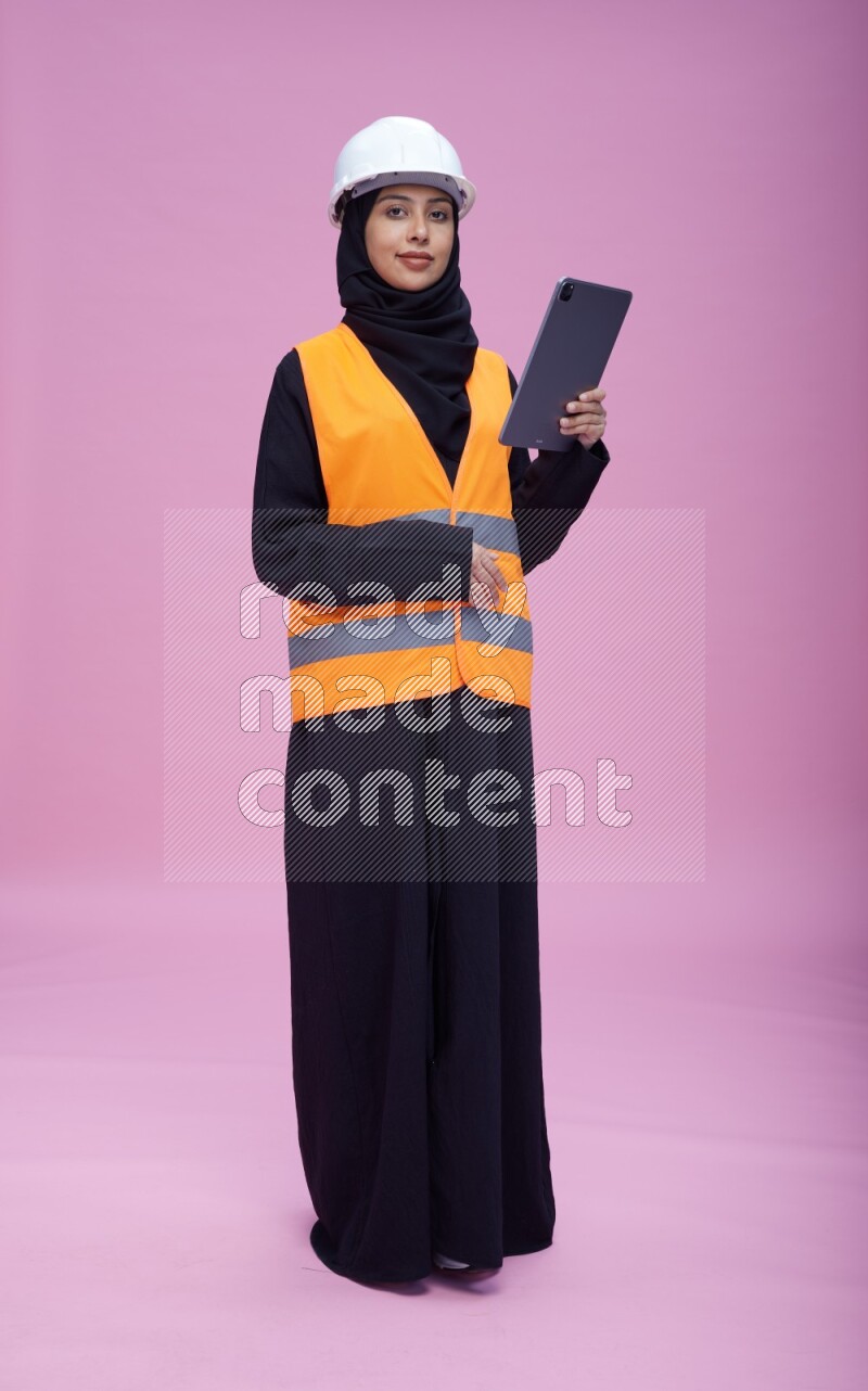 Saudi woman wearing Abaya with engineer vest and helmet standing working on tablet on pink background