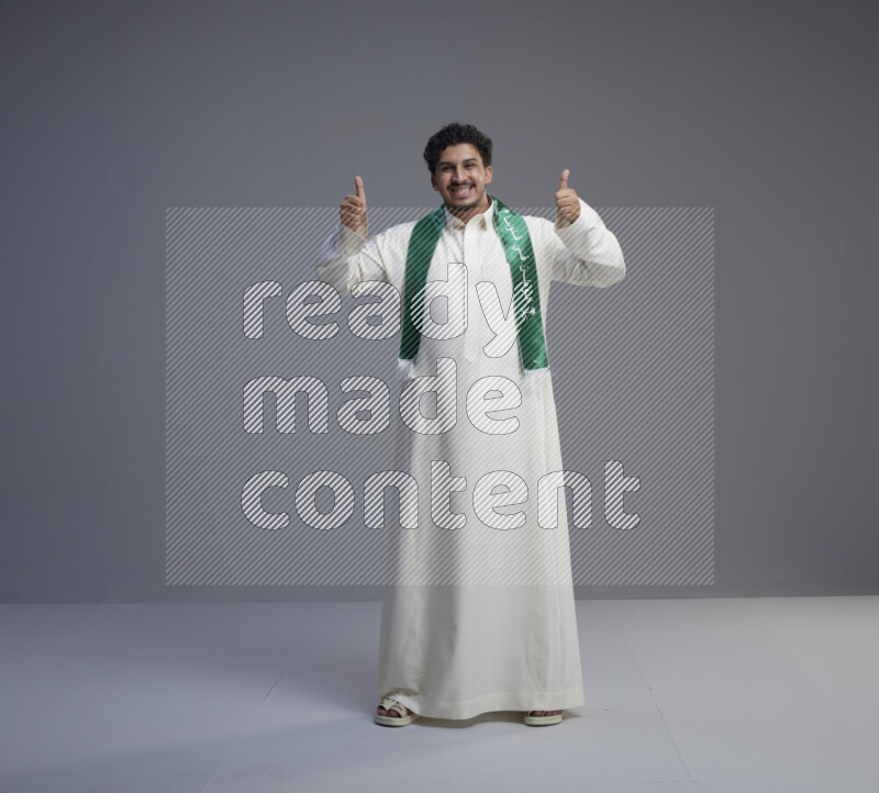 A Saudi man standing wearing thob and Saudi flag scarf on gray background