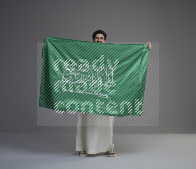 A Saudi man standing wearing thob holding big Saudi flag on gray background