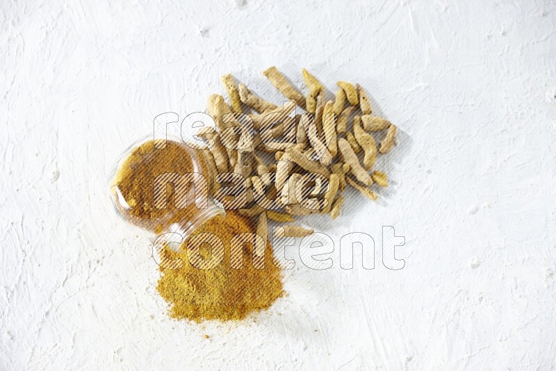 A flipped glass spice jar full of turmeric powder and powder spilled out of it with dried whole fingers on textured white flooring