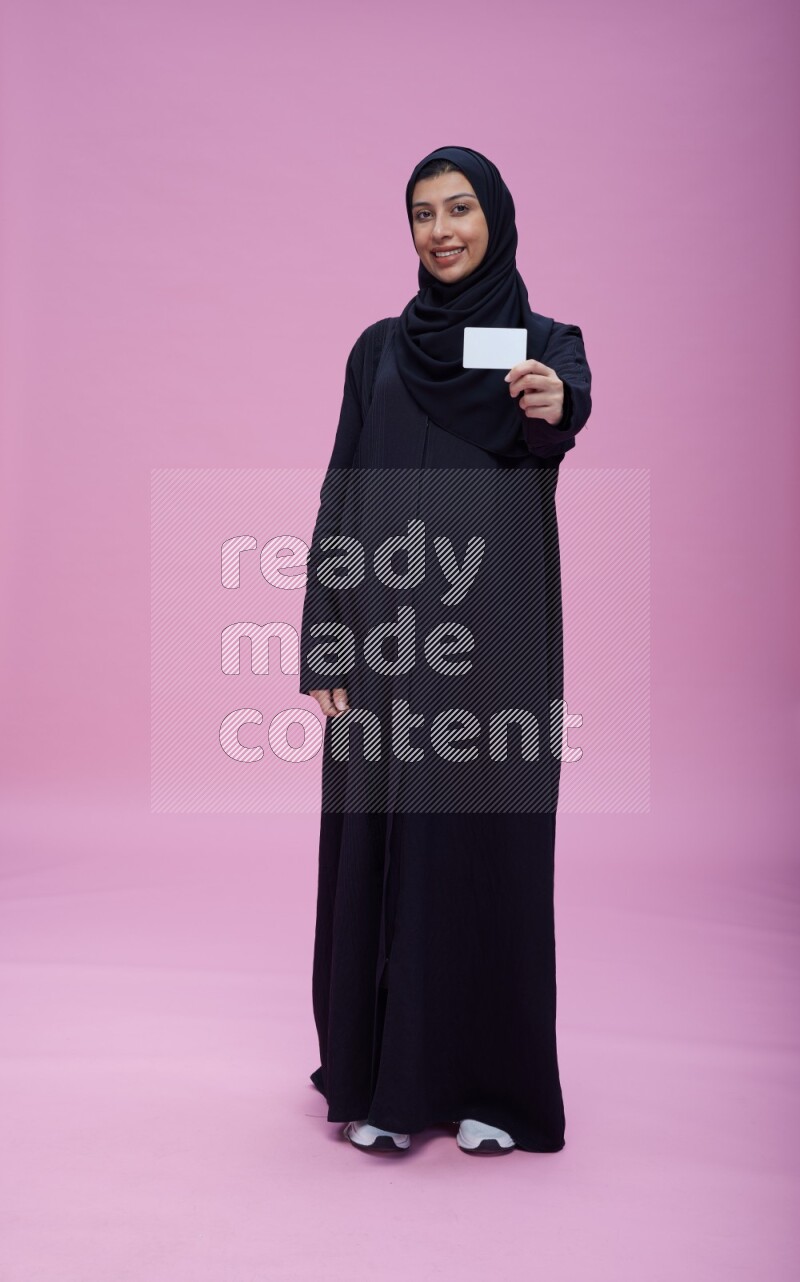Saudi woman wearing Abaya standing holding ATM card on pink background