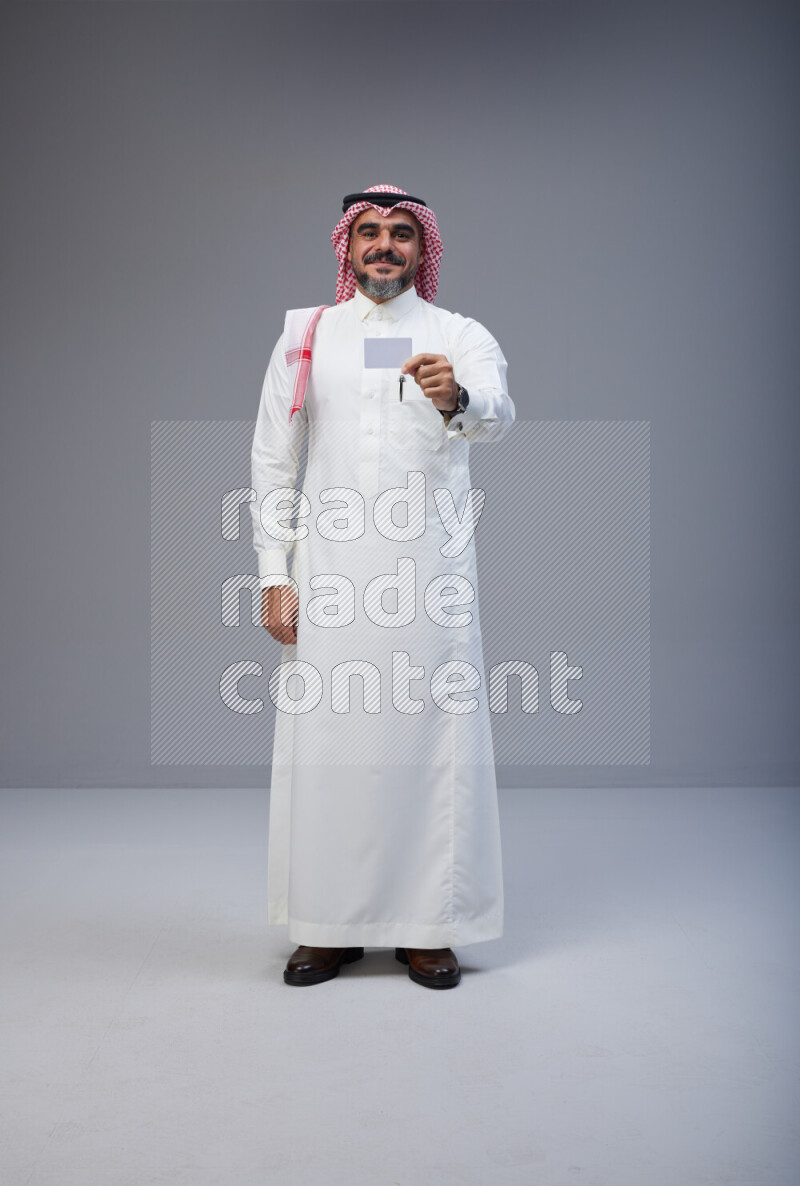 Saudi man Wearing Thob and red Shomag standing holding ATM card on Gray background