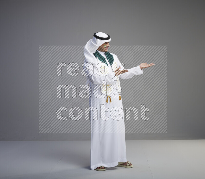 A Saudi man standing wearing thob and white shomag with face painting and Saudi flag scarf on gray background