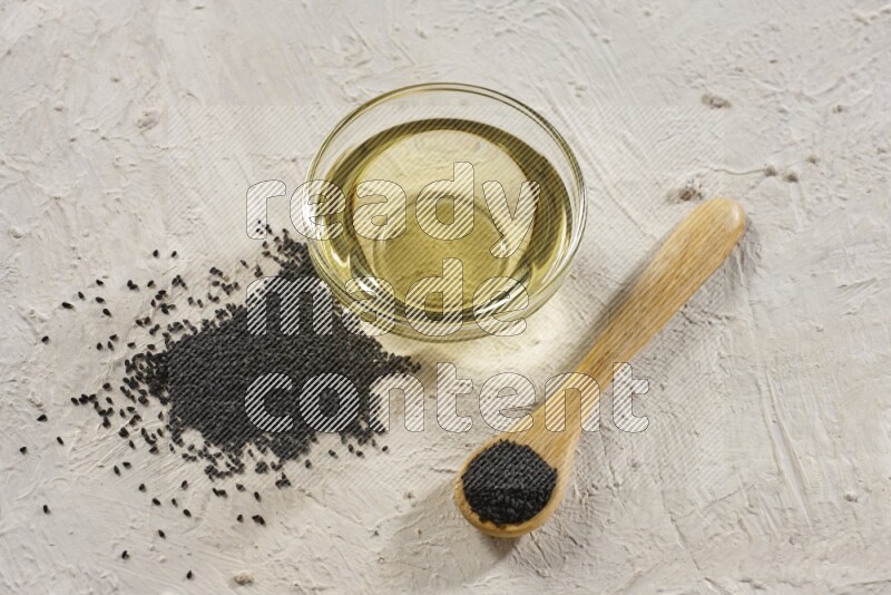 A glass bowl full of black seeds oil and wooden spoon full of black seeds with seeds spread on a textured white flooring