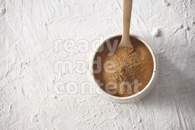 Ceramic beige bowl full of cinnamon powder with a wooden spoon on a textured white background