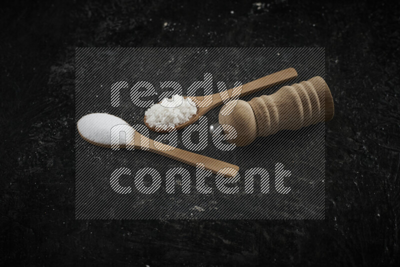 2 wooden spoons filled with fine and coarse salt with wooden grinder beside them on black background