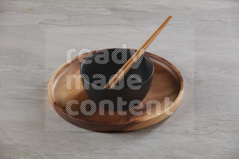 black ceramic round bowl on top of brown wood round plate and wood chopsticks, on grey textured countertop