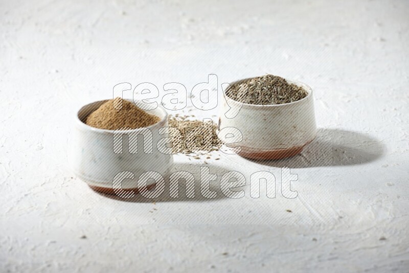 2 beige bowls full of cumin seeds and powder with spilled powder and seeds on textured white flooring