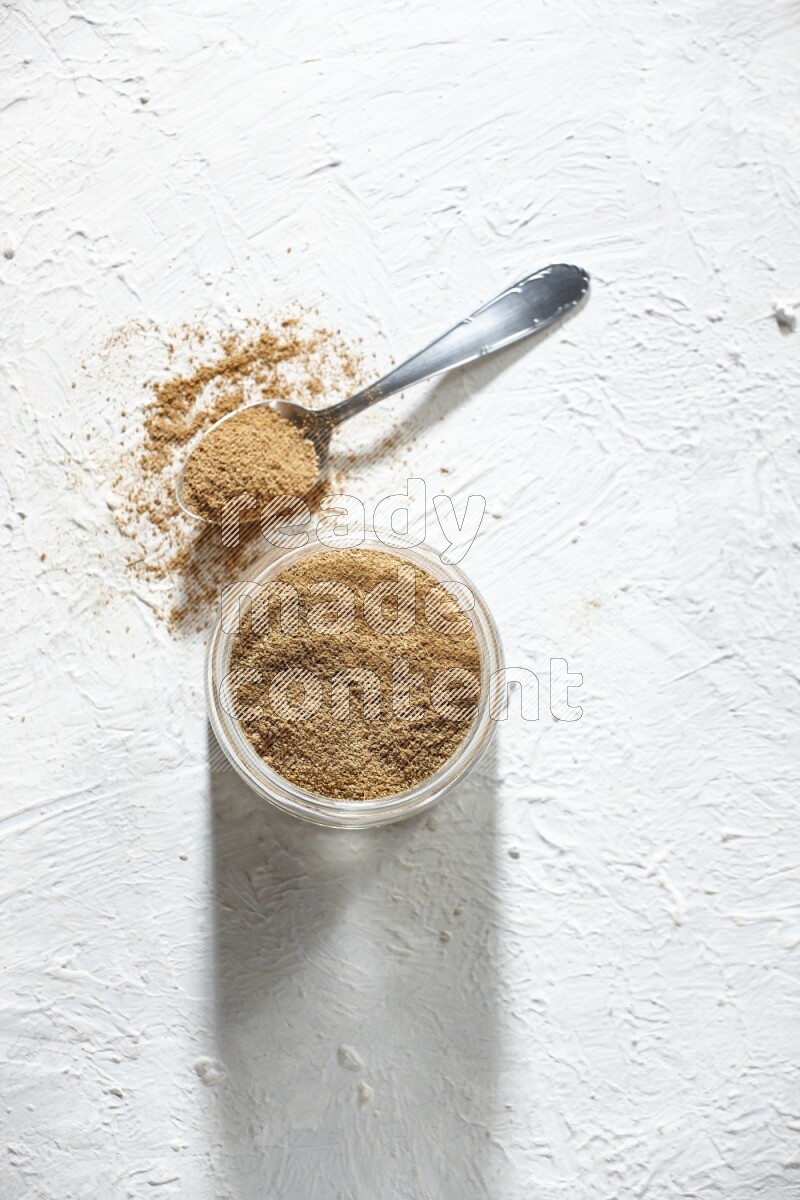 A glass jar and a metal spoon full of cumin powder on textured white flooring