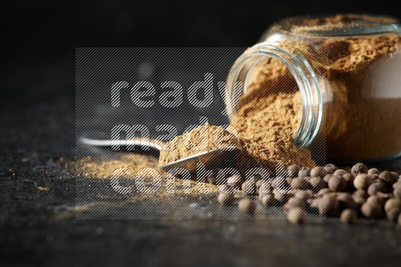 A flipped glass spice jar and metal spoon full of allspice powder and powder spilled out of it with whole balls on a textured black flooring