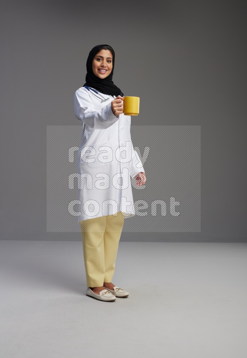 Saudi woman wearing lab coat with stethoscope standing holding mug on Gray background