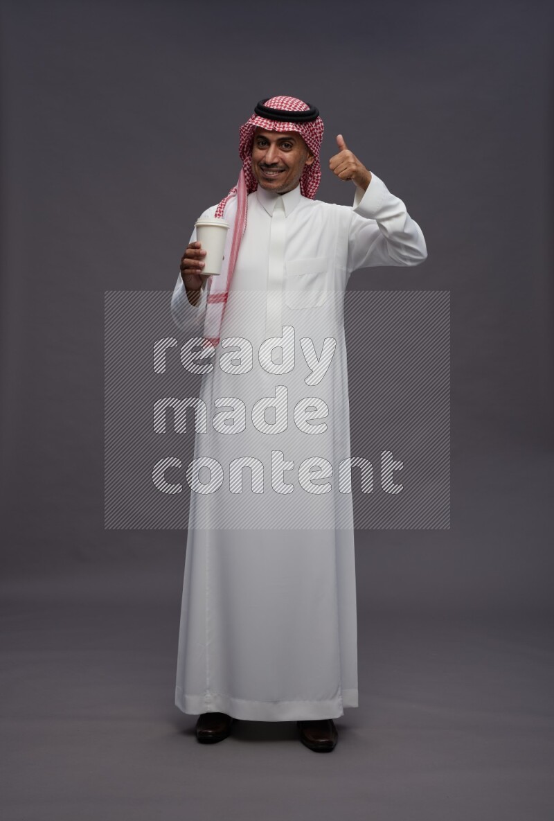 Saudi man wearing thob and shomag standing holding paper cup on gray background