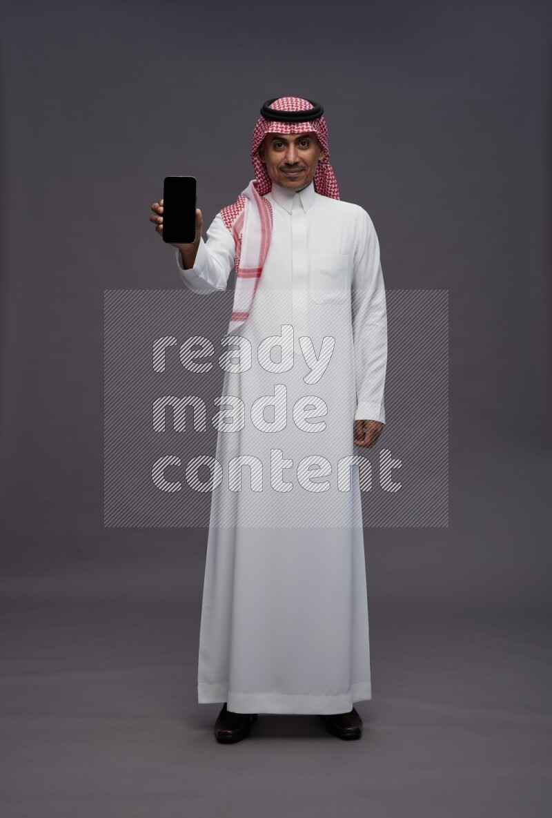 Saudi man wearing thob and shomag standing showing phone to camera on gray background