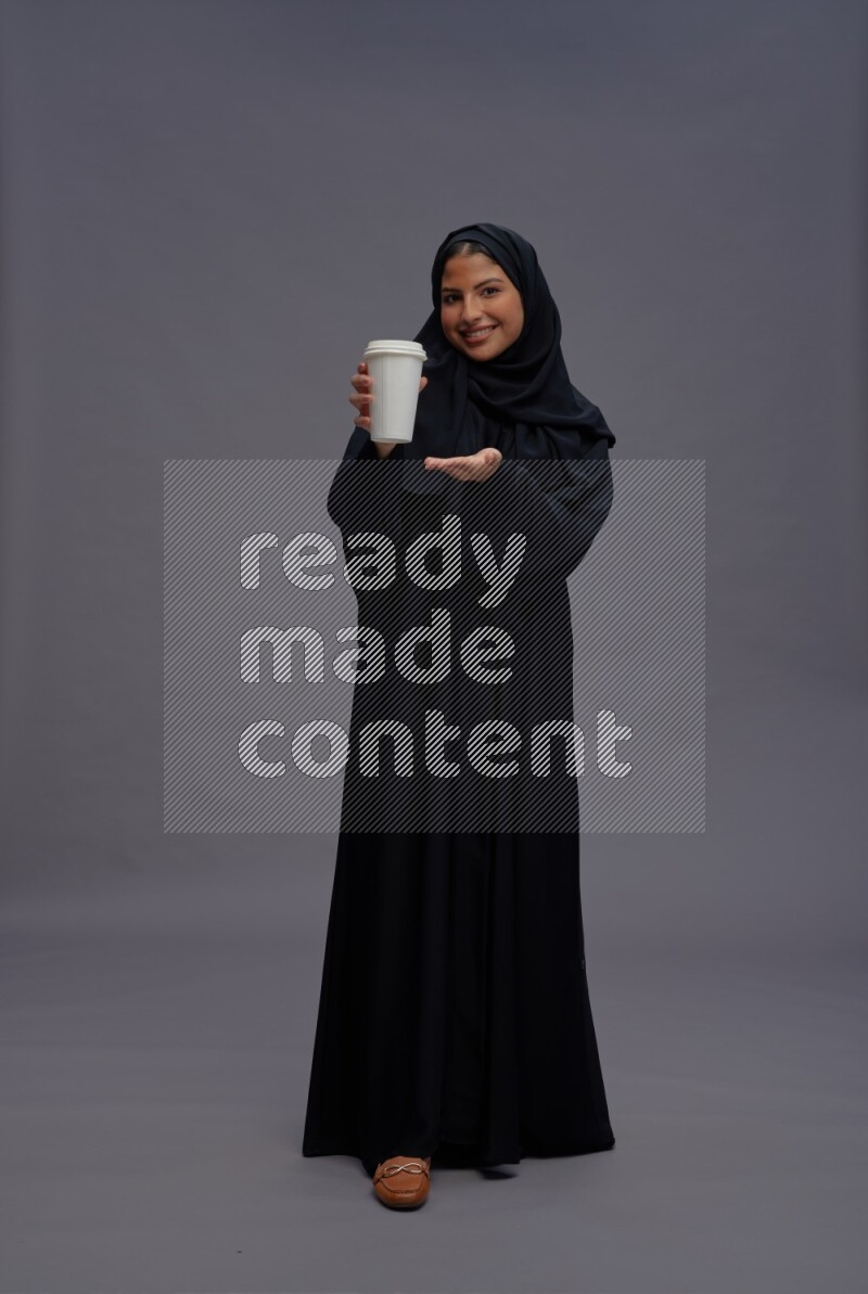 Saudi woman wearing Abaya standing holding paper cup on gray background