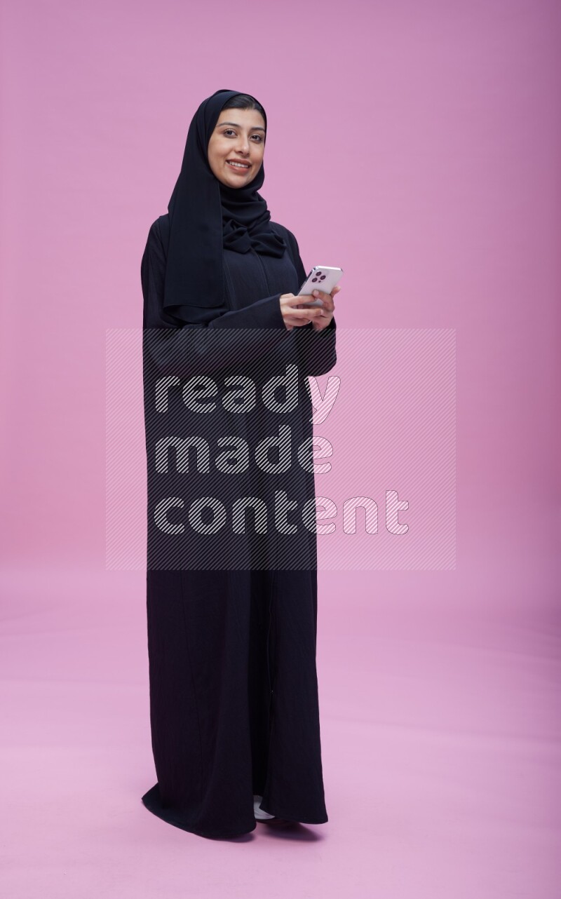 Saudi woman wearing Abaya standing texting on phone on pink background