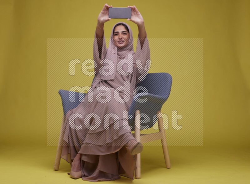 A woman Showing Her Phone Screen Setting  on a Yellow Background wearing Brown Abaya with Hijab