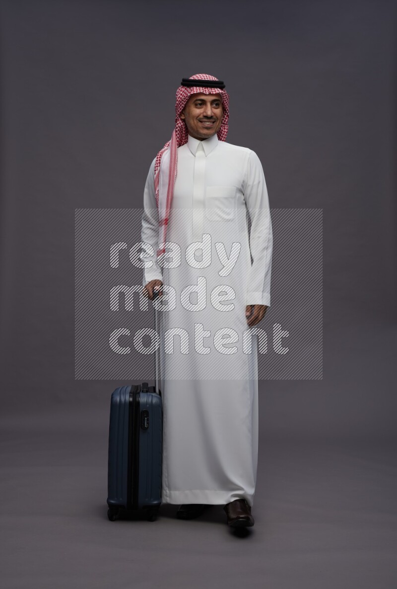 Saudi man wearing thob and shomag standing holding bag on gray background
