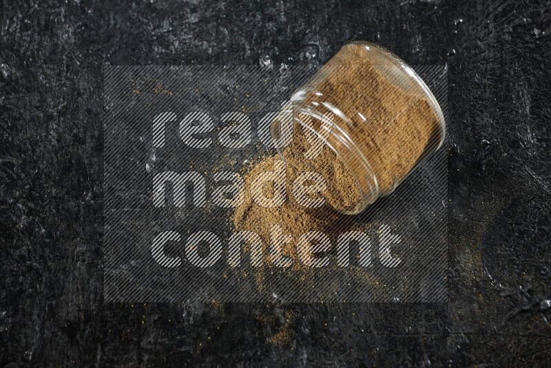 A flipped glass jar full of cumin powder and powder fell out on a textured black flooring