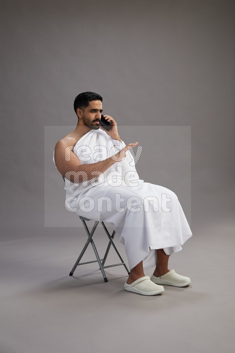 A man wearing Ehram sitting on chair talking on the phone on gray background