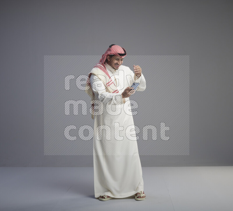 A  Saudi man standing wearing thob and red shomag texting on phone on gray background