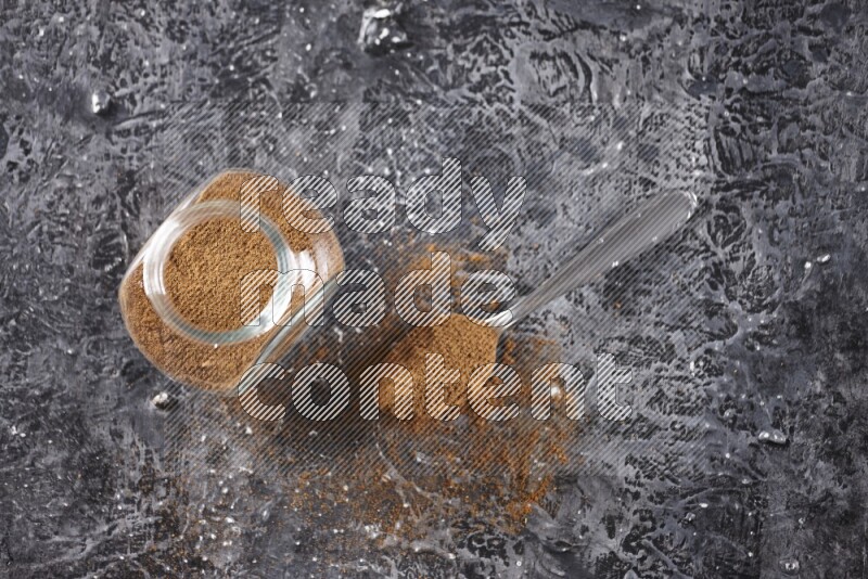 Herbal glass jar full of cinnamon powder and a metal spoon full of powder on textured black background
