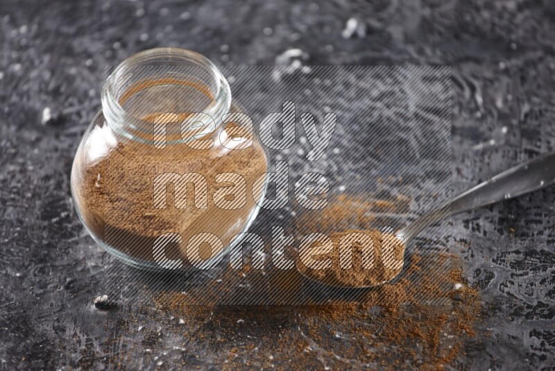 Herbal glass jar full of cinnamon powder and a metal spoon full of powder on textured black background