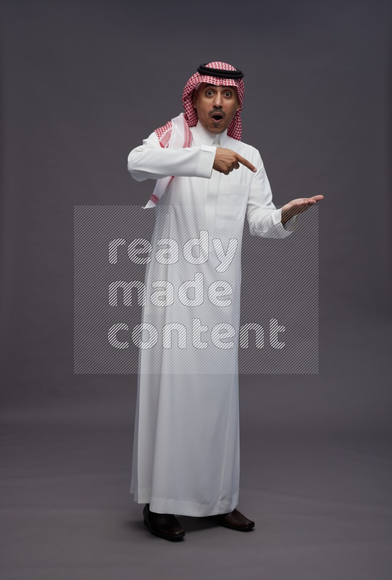 Saudi man wearing thob and shomag standing interacting with the camera on gray background