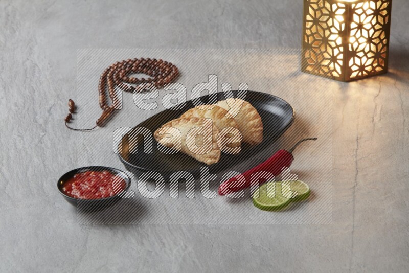 Three fried sambosas in an oval shaped black plate and a red sauce in a black round ramekin with a brown misbaha and a golden lantern on the side on a gray background